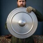 Brushed steel round shield with riveted boss plate held up by a gloved hand in front of a bearded man