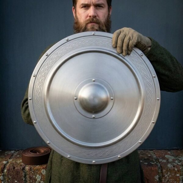 Brushed steel round shield with riveted boss plate held up by a gloved hand in front of a bearded man