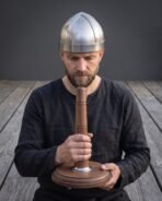 Brushed steel spangenhelm cap worn by a bearded man in black who holds the runed walnut display stand in both hands