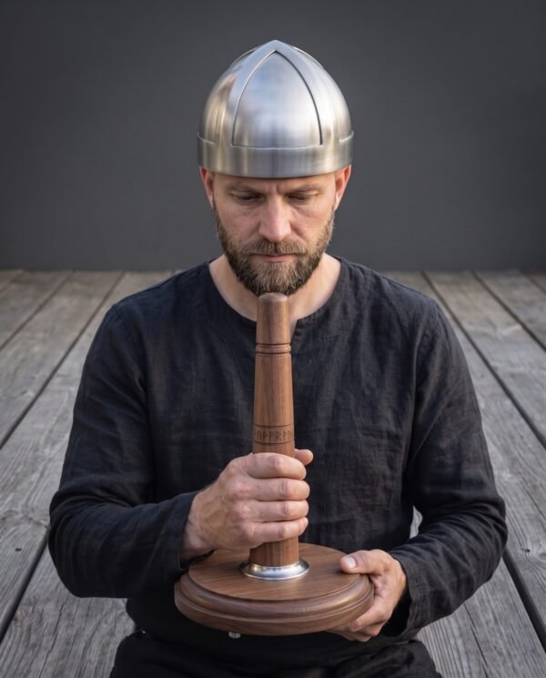 Brushed steel spangenhelm cap worn by a bearded man in black who holds the runed walnut display stand in both hands
