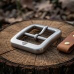 Brushed steel square belt buckle with Valknut stamp, angled close up on a cut oak round beside a tan leather tab