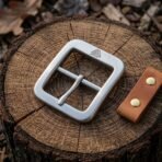 Brushed steel Viking belt buckle with centre bar and shaped tongue resting on a sawn log next to a leather snap tab