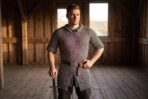 Butted mild steel chainmail hauberk with short sleeves worn with a brown leather belt by a man standing inside a timber barn