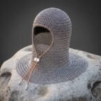 Butted steel chainmail coif with leather bound face opening, tan drawstring and wide shoulder mantle resting on a pale boulder