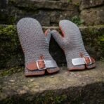 Pair of butted steel chainmail mittens with tan leather cuffs and knotwork buckle plates standing on a mossy stone step
