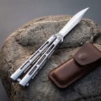 Butterfly knife with open channel handles and clip point blade angled across a rock, brown leather pouch in front