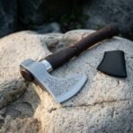 Carbon steel bearded axe head close up with etched runes and staves, steel collar and dark haft next to a black leather cover