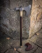 Carbon steel bearded axe with etched knotwork head, brass collar and dark haft leaning on a stone wall beside its leather mask