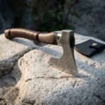 Carbon steel bearded axe with rune etched head and leather cord wrapped dark wood haft on a rock beside a black edge cover