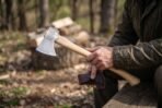 Carbon steel felling axe with chased knotwork cheek and curved pale wood haft held by a seated man with a leather blade mask