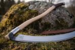 Carbon steel scythe blade close up with etched triquetra, steel socket ferrule and pale ash haft on moss beside a leather cover