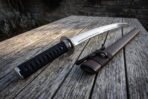 Carbon steel wakizashi grip and blade close up with round steel guard beside the cord bound brown leather saya on planks
