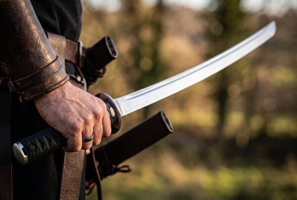 Carbon steel wakizashi held raised in one hand beside a belt carried brown leather saya with blurred trees behind