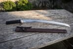 Carbon steel wakizashi with black ribbed grip and steel guard lying above its dark brown leather saya on a weathered table