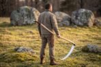 Carbon steel war scythe with long pale ash haft carried by a man standing in a meadow among mossy boulders