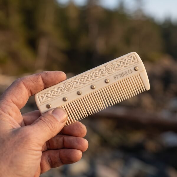 Carved bone comb with knotwork band and rune line held between fingers against a blurred forest shore