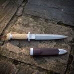 Carved bone hilted Damascus Celtic dagger with Elder Futhark runes etched at the ricasso lying on grey paving beside a burgundy leather sheath