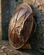 Carved hardwood round shield with eagle and six spoke relief, bronze toned rim and leather arm straps against a stone wall
