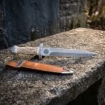 Celtic dagger with a carved bone handle and folded Damascus blade resting on a granite ledge above a tan leather sheath with polished steel throat and chape