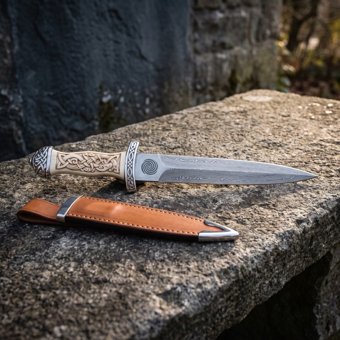 Celtic dagger with a carved bone handle and folded Damascus blade resting on a granite ledge above a tan leather sheath Celtic dagger with a carved bone handle and folded Damascus blade resting on a granite ledge above a tan leather sheath with polished steel throat and chape