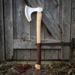 Celtic knotwork bearded axe with leather wrapped ash haft leaning on a weathered barn door, leather edge sheath below