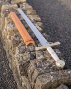 Celtic knotwork sword laid along the top of a dry stone wall beside its tan leather scabbard, white leather grip and disc pommel bright in the evening light