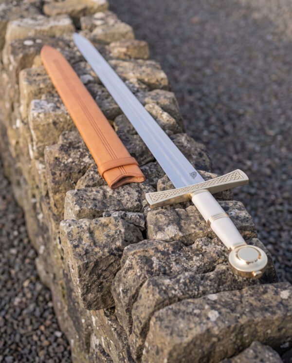 Celtic knotwork sword laid along the top of a dry stone wall beside its tan leather scabbard, white leather grip and disc pommel bright in the evening light