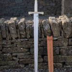 Celtic knotwork sword standing point down in front of a dry stone wall, bright 1095 blade below a brass toned engraved guard with its tan leather scabbard alongside