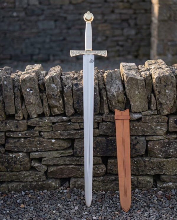 Celtic knotwork sword standing point down in front of a dry stone wall, bright 1095 blade below a brass toned engraved guard with its tan leather scabbard alongside