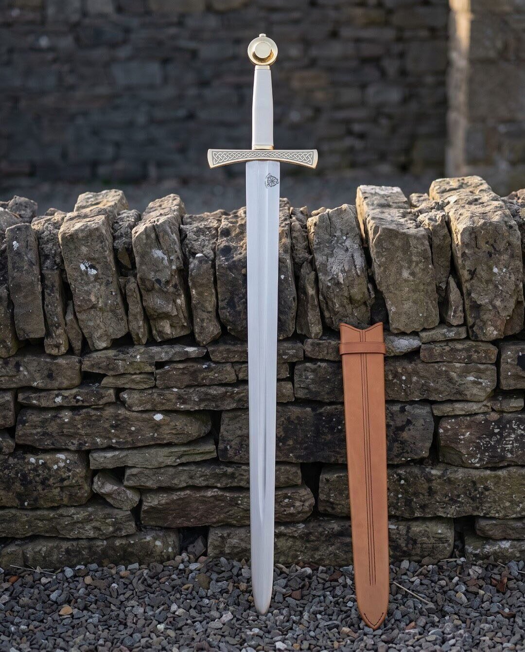 Celtic knotwork sword standing point down in front of a dry stone wall, bright 1095 blade below a brass toned engraved g Celtic knotwork sword standing point down in front of a dry stone wall, bright 1095 blade below a brass toned engraved guard with its tan leather scabbard alongside