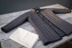 Charcoal cotton canvas padded gambeson laid at an angle on a weathered wooden table, showing the quilted channels and brass grommet lacing