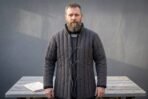 Charcoal cotton canvas padded gambeson with vertical channel quilting, standing collar and front cord lacing worn by a bearded man