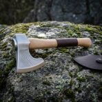 Chevron etched Viking axe in satin 1095 steel with a hammer poll and leather wrapped white ash haft resting on a moss and lichen covered granite boulder