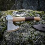 Chevron etched Viking axe in satin 1095 steel with a hammer poll and leather wrapped white ash haft resting on a moss and lichen covered granite boulder