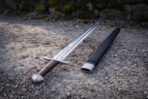 Chevron fuller viking sword in polished 1095 carbon steel with a lobed pommel and brown leather grip, lying on gravel beside its black leather scabbard by a mossy wall