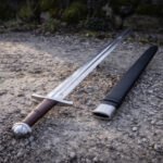 Chevron fuller viking sword in polished 1095 carbon steel with a lobed pommel and brown leather grip, lying on gravel beside its black leather scabbard by a mossy wall