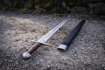 Chevron fuller viking sword photographed from the pommel end on gravel, the etched herringbone band and struck sun mark visible in the fuller beside the black leather scabbard