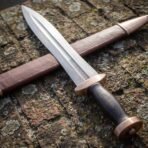 Close angled view of a Damascus gladius blade with Greek key border, bronze guard and dark grip resting across its scabbard