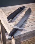 Close angled view of a knotwork damascus falchion lying on a grey wooden table beside its tooled black leather sheath, showing the knot carved guard and cap pommel