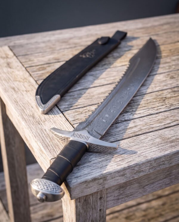 Close angled view of a knotwork damascus falchion lying on a grey wooden table beside its tooled black leather sheath, showing the knot carved guard and cap pommel