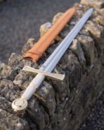 Close detail of a celtic knotwork sword on limestone, the raised plaitwork band engraved along the brass toned guard above a white leather wrapped grip