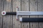 Close overhead detail of a jewelled arming sword showing the etched Norse plaitwork band on the blade, the maker roundel and red set stones in the straight steel guard