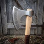Close up of a bearded axe head with Celtic knotwork engraving along the cheek on a pale ash haft against an old door