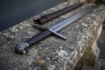 Close up of a cast wolf head pommel and dark ringed grip on a Damascus arming sword resting on a lichen covered bench