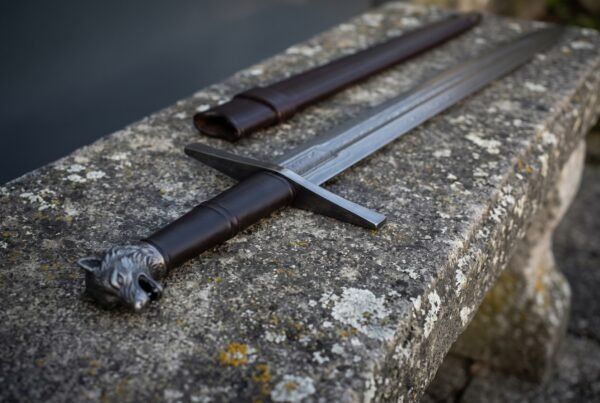 Close up of a cast wolf head pommel and dark ringed grip on a Damascus arming sword resting on a lichen covered bench