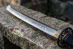 Close up of a curved sword blade with wavy temper line, engraved brass collar and black openwork guard on rough granite