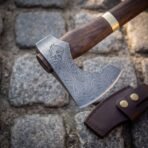 Close up of a Damascus bearded axe head with etched wolf, walnut haft and brass collar above a brown leather edge sheath