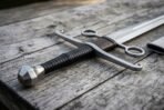 Close up of a hand and a half sword hilt with faceted steel pommel, black spiral grip and ring guard on weathered planks