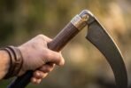Close up of a hand gripping the hardwood haft of a Damascus war scythe below an engraved brass collar and spiral