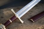 Close up of a knightly sword hilt with brass guard, oxblood leather grip and knotwork etched fuller on stone